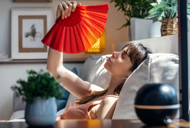 Una donna seduta su un divano usa un ventilatore per rinfrescarsi